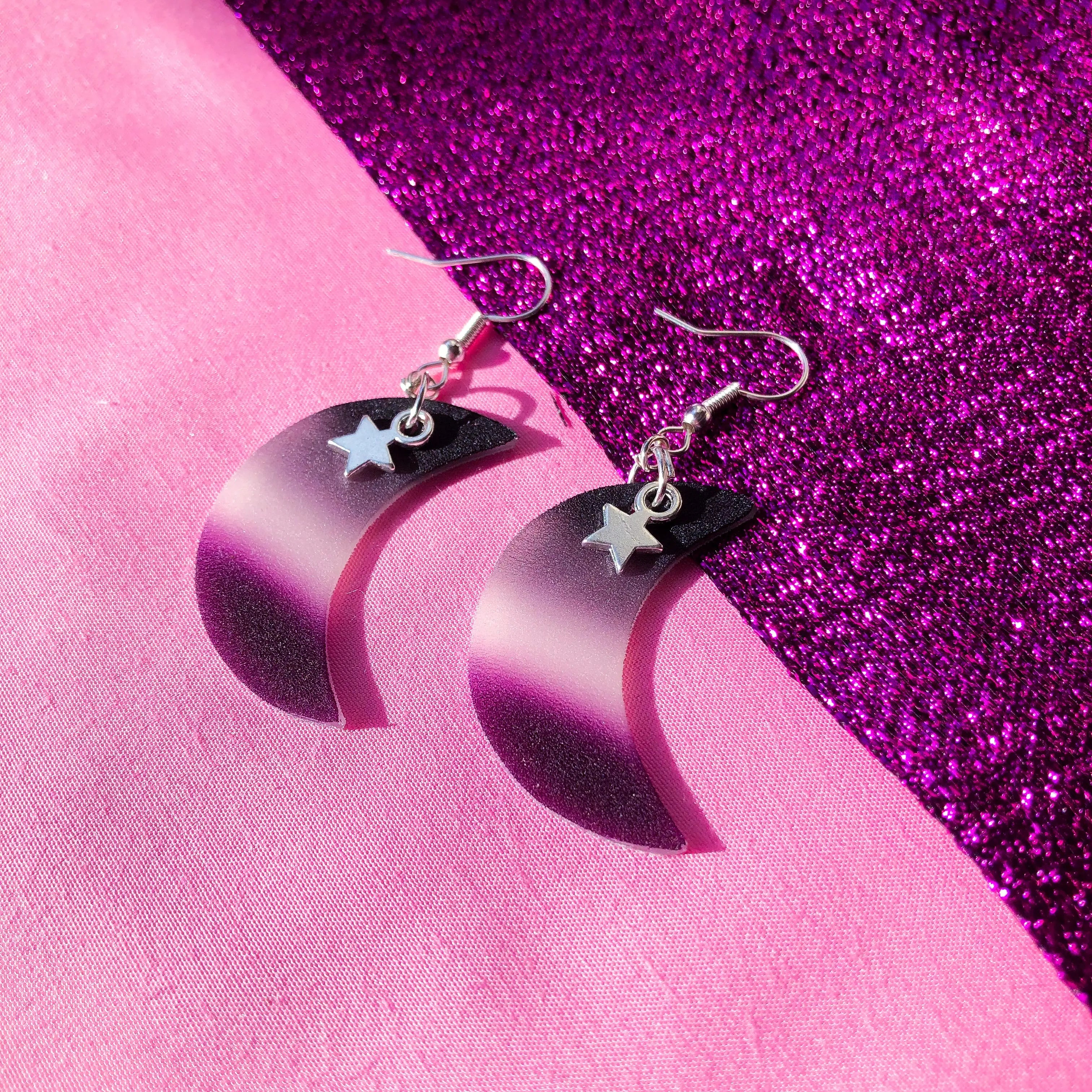 Shrinky dink earrings in the shape of a crescent moon. The colour of the charm is the asexual pride flag with a small gold star charm hanging at the top.